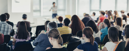 A group of people seated in chairs, attentively listening to a speaker at the front of the room.