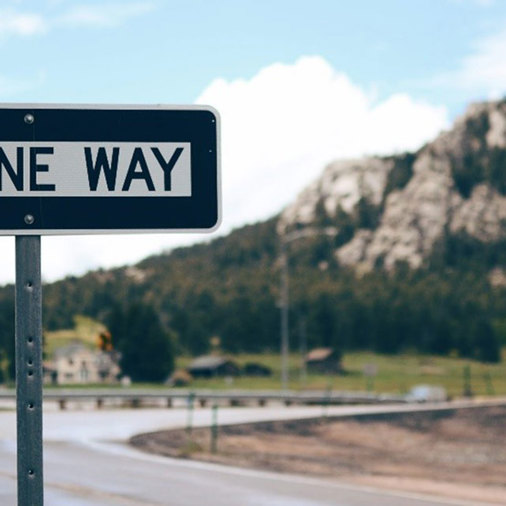 Road sign indicating one-way traffic, set against a scenic mountain view in the background.