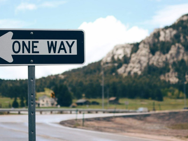 Road sign indicating one-way traffic, set against a scenic mountain view in the background.