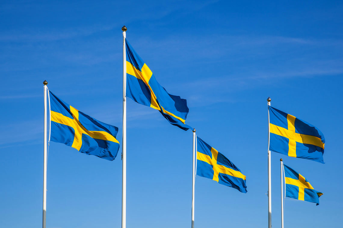 Swedish flags fluttering in the wind against a clear blue sky.