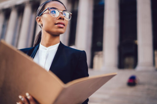A businesswoman wearing glasses holds a folder, looking confidently at the camera in a professional setting.