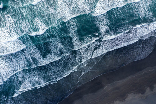 Aerial view of ocean waves crashing onto a dark sandy beach, creating white foamy patterns.