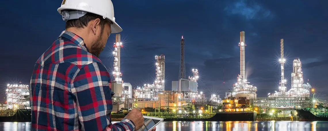 A man in a hard hat and plaid shirt observes a large refinery in the background, showcasing an industrial setting.