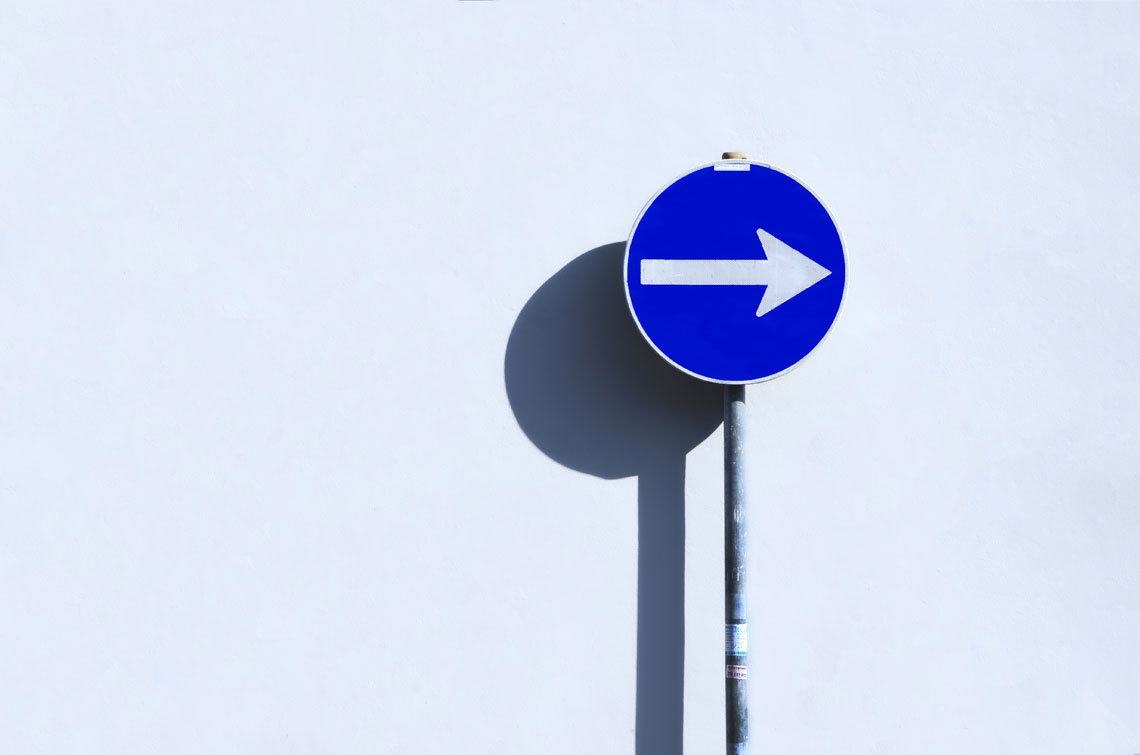 A blue and white directional sign with an arrow indicating the way to the road ahead.