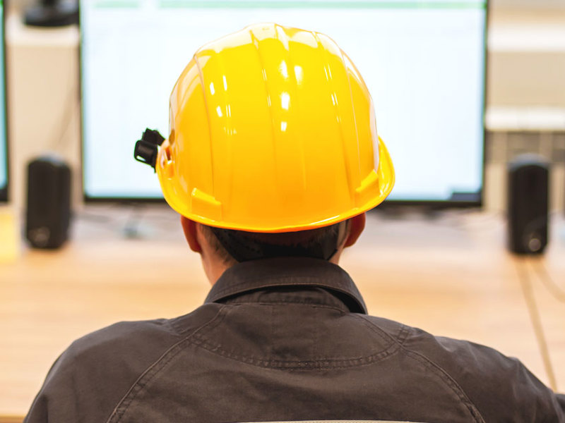 A man in a hard hat sits at a desk, focused on multiple computer monitors displaying various data and information.